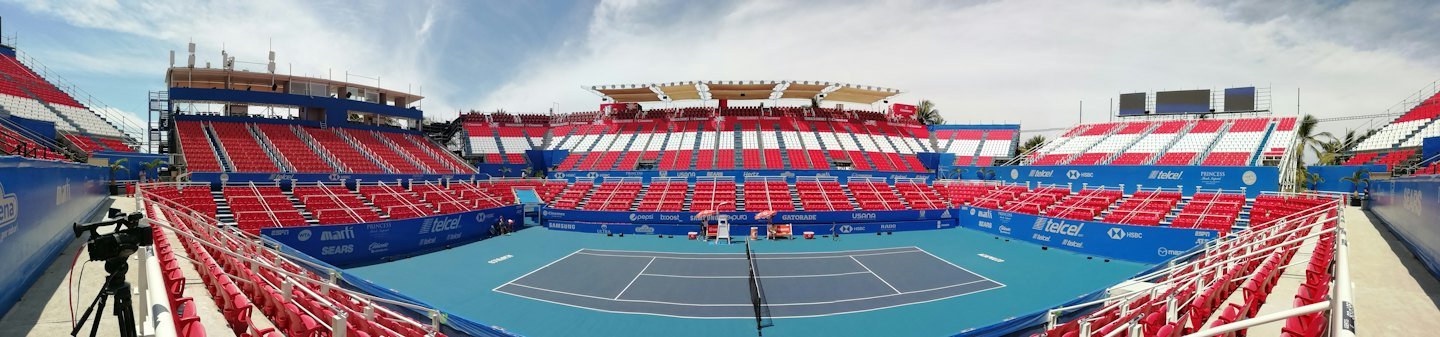 Empty tennis stadium with blue courts and tiered spectator seating.